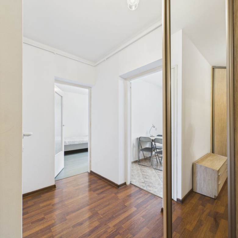 Hallway with white walls and dark wood floor, open doors to a bedroom on the left and a tiled room with chairs on the right; a mirrored wardrobe on the right.