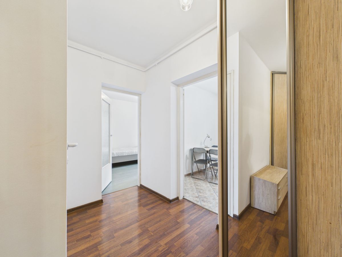 Hallway with white walls and dark wood floor, open doors to a bedroom on the left and a tiled room with chairs on the right; a mirrored wardrobe on the right.
