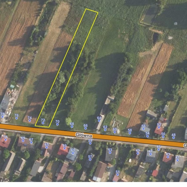 Aerial view of a long narrow plot outlined in yellow along Główna street, between trees and houses.
