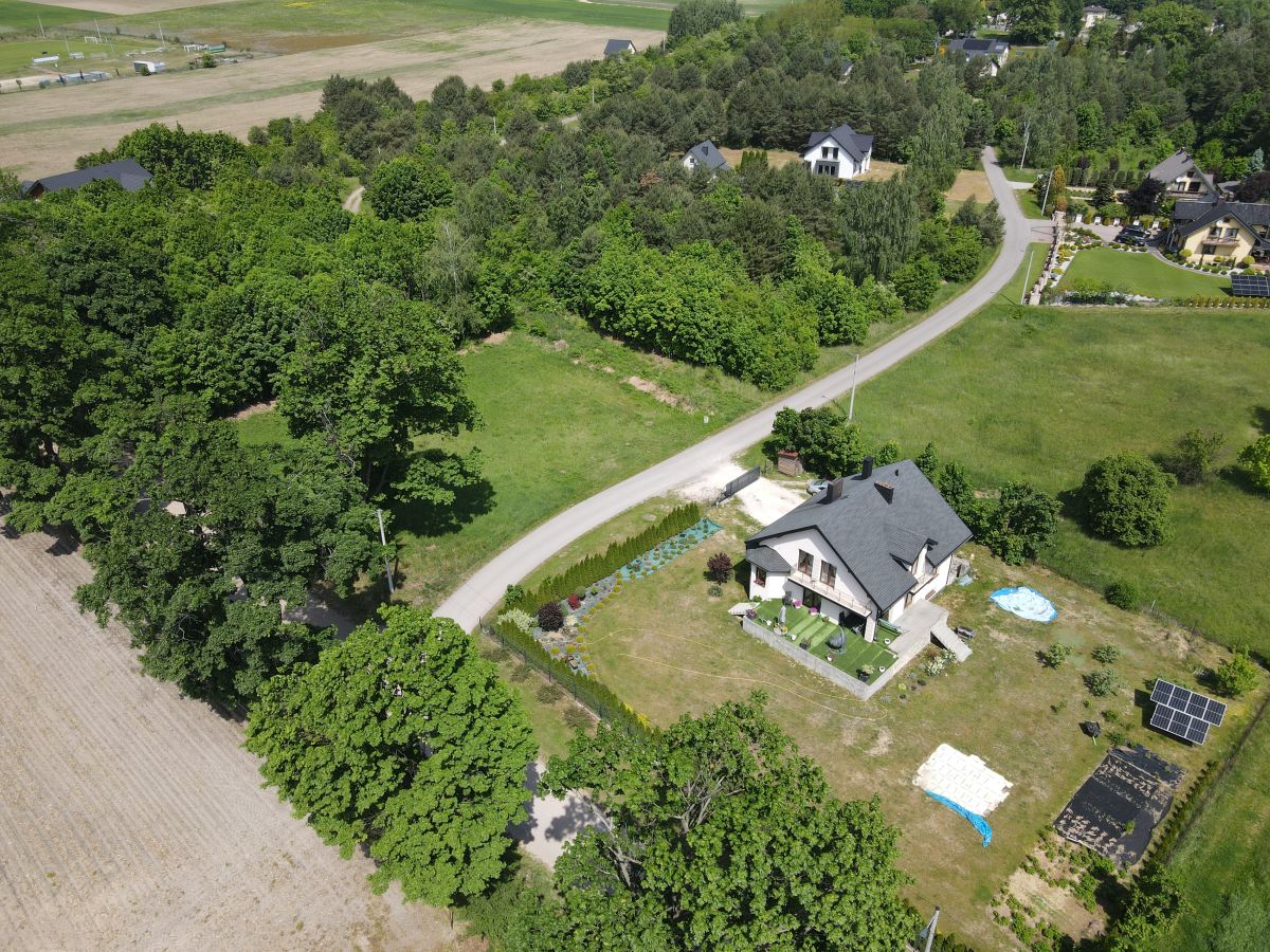 Aerial view of a two-story house with a gray roof, green lawn, and a curved road through a rural area surrounded by trees.