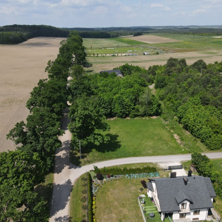 Aerial view of a two-story house with a gray roof at bottom right, surrounded by trees, a gravel road, and open fields beyond.