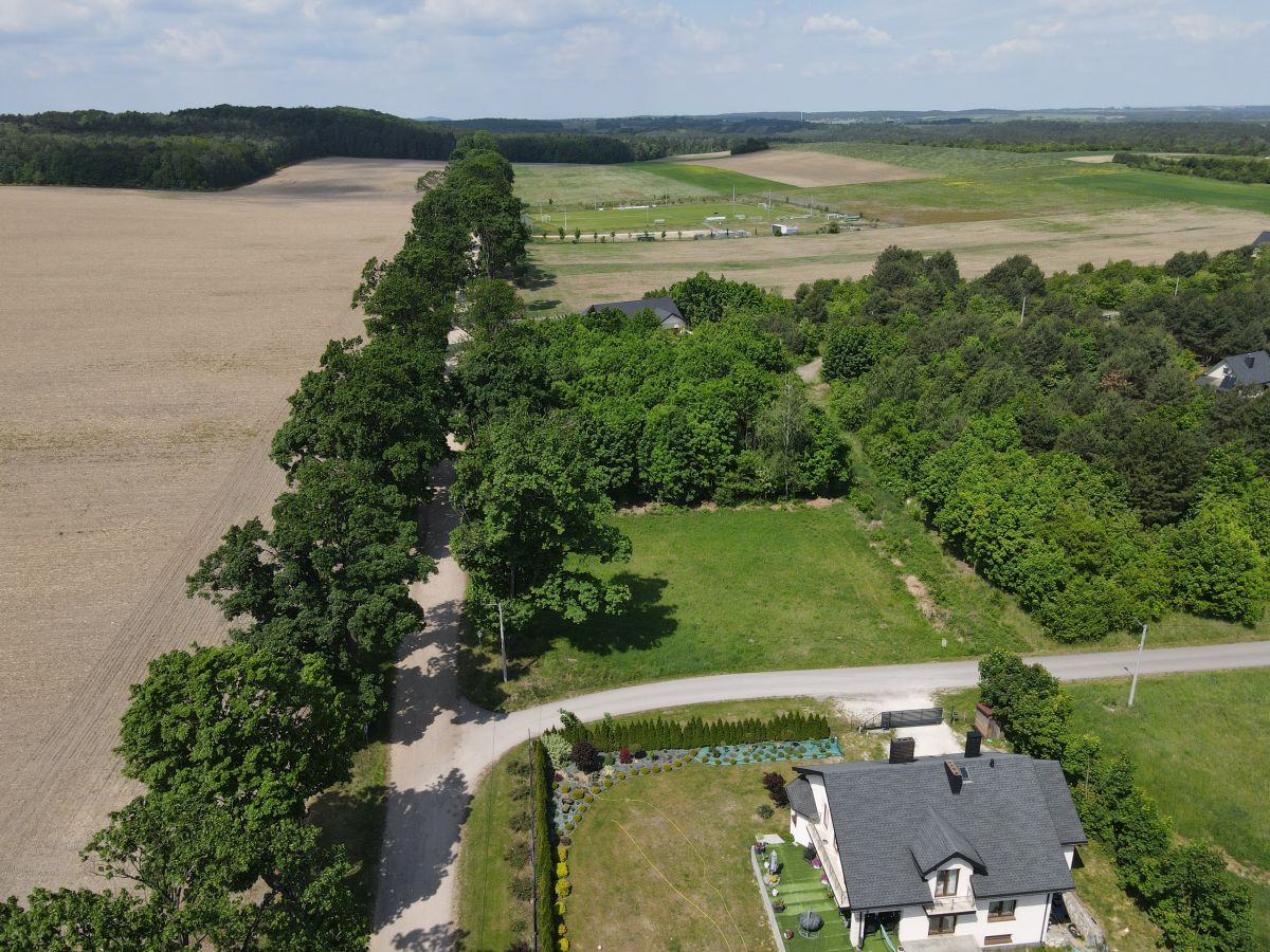 Aerial view of a two-story house with a gray roof at bottom right, surrounded by trees, a gravel road, and open fields beyond.