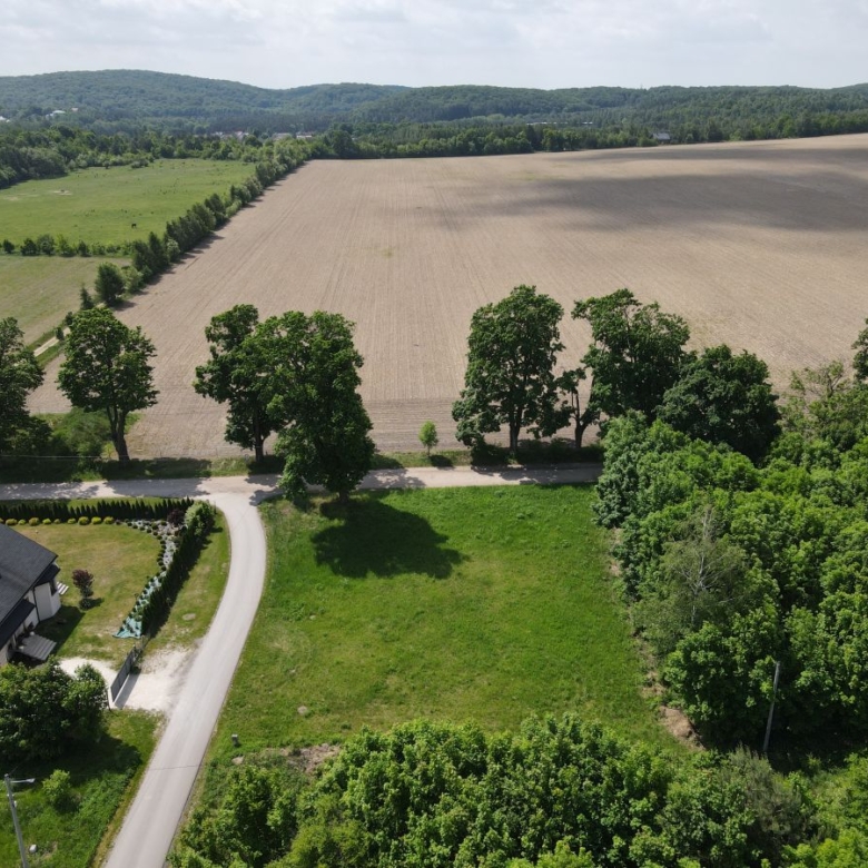 Aerial view of countryside: a curved road beside a white house with a lawn, lined by trees, leading to a large plowed field and distant hills.