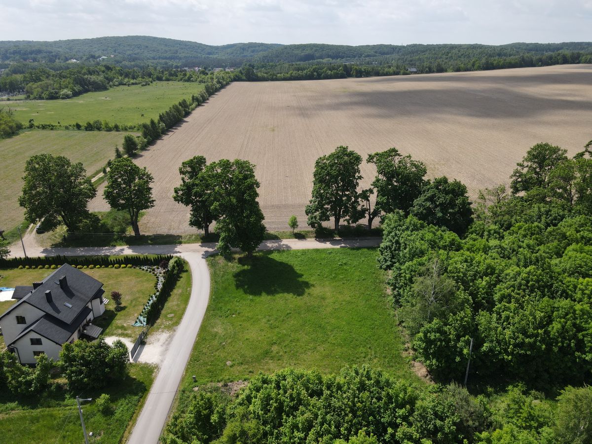 Aerial view of countryside: a curved road beside a white house with a lawn, lined by trees, leading to a large plowed field and distant hills.