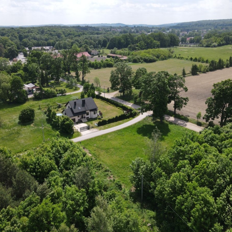 Aerial view of a two-story house with dark roof along a curved road, surrounded by trees and green fields.