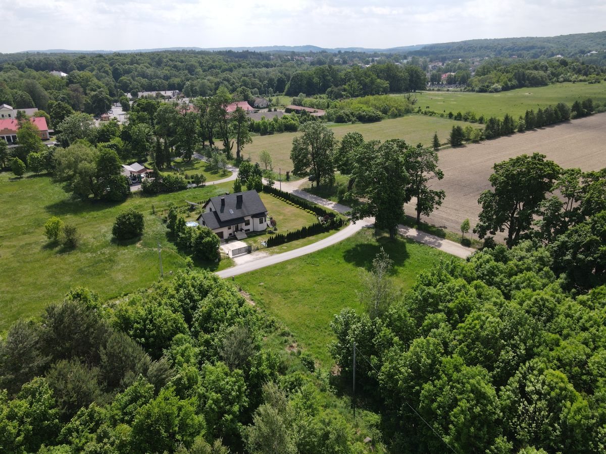 Aerial view of a two-story house with dark roof along a curved road, surrounded by trees and green fields.