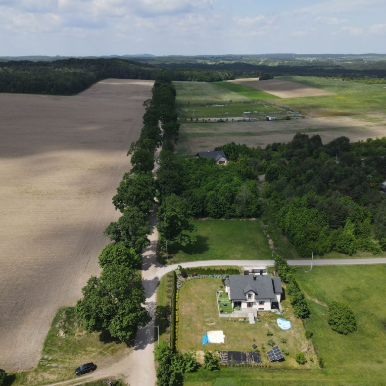 Aerial view of a rural property: a house with a fenced yard, solar panels, a small pool, and surrounding fields and trees.