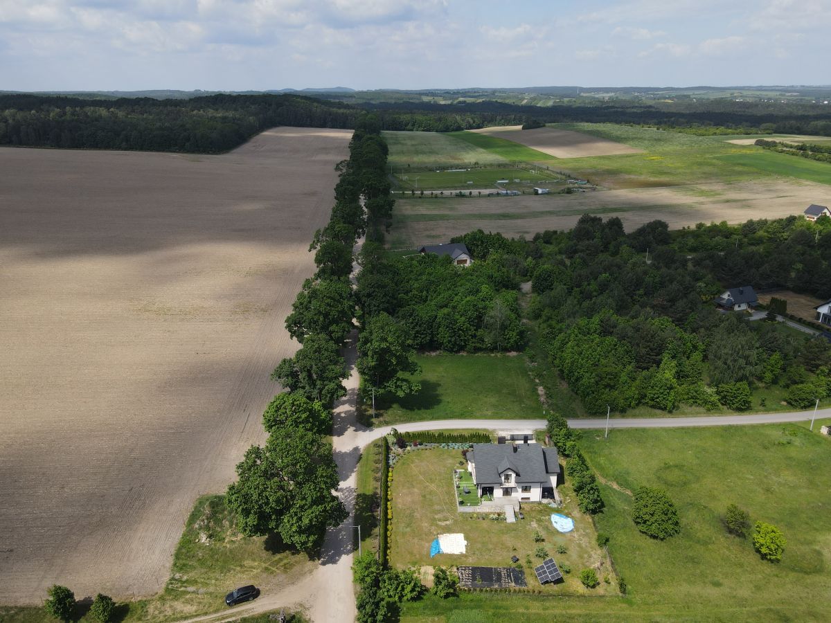 Aerial view of a rural property: a house with a fenced yard, solar panels, a small pool, and surrounding fields and trees.
