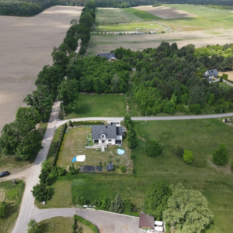 Aerial view of a rural property with a grey-roof house, a fenced yard, solar panels and a blue pool in the yard. Nearby fields and trees border the scene.
