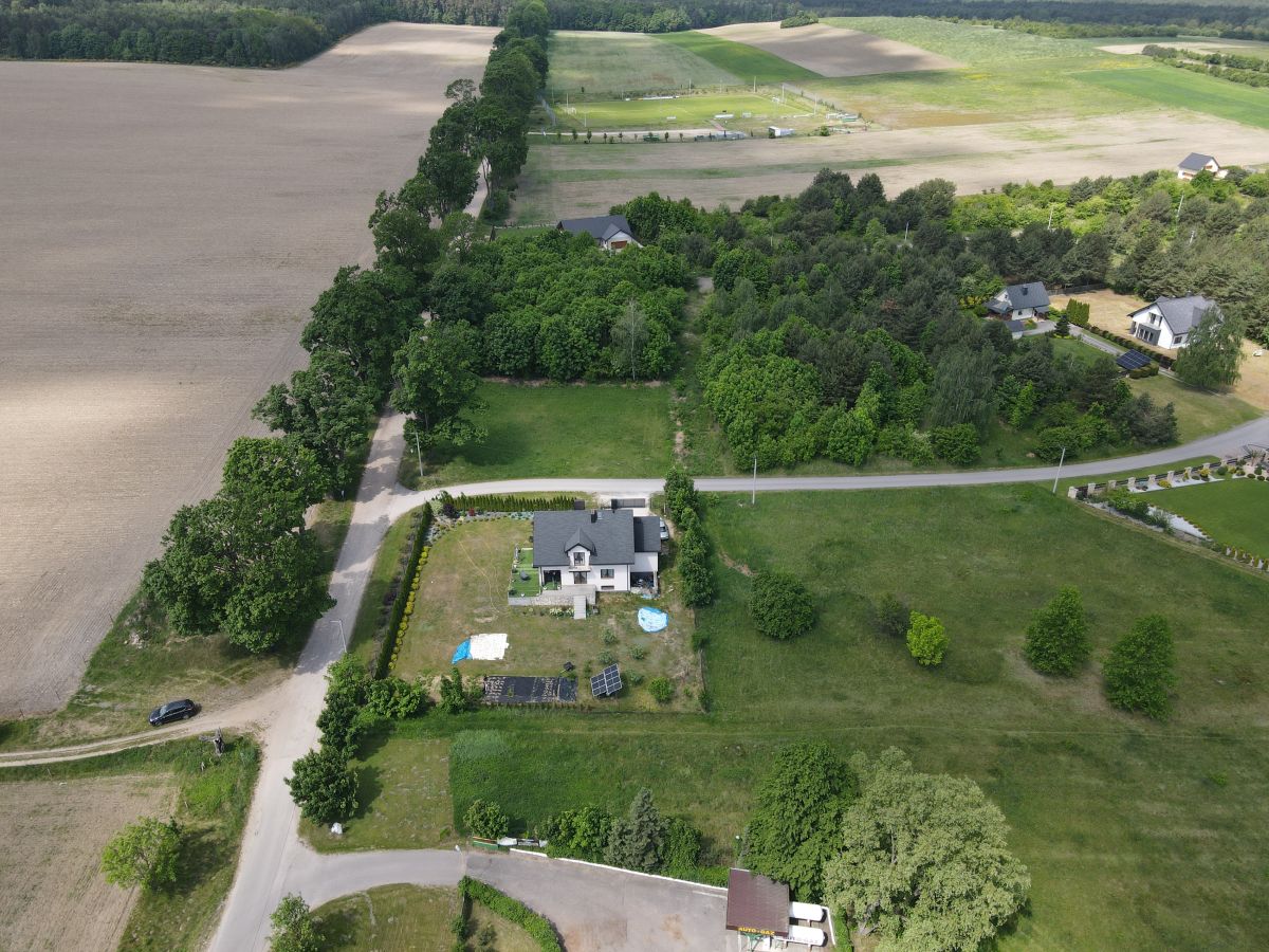 Aerial view of a rural property with a grey-roof house, a fenced yard, solar panels and a blue pool in the yard. Nearby fields and trees border the scene.