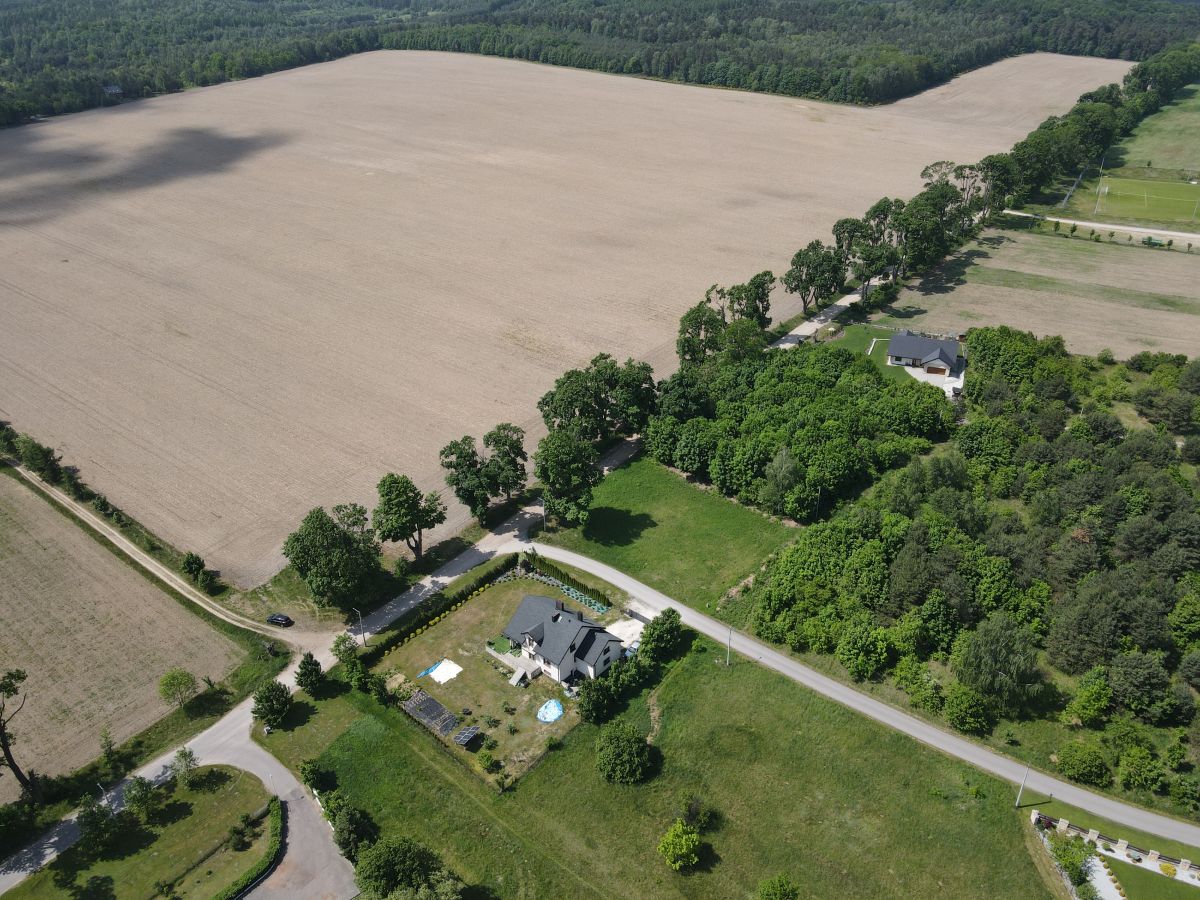 Aerial view of a rural landscape showing a large harvested field bordered by a line of trees and a winding road with houses and green yards nearby.