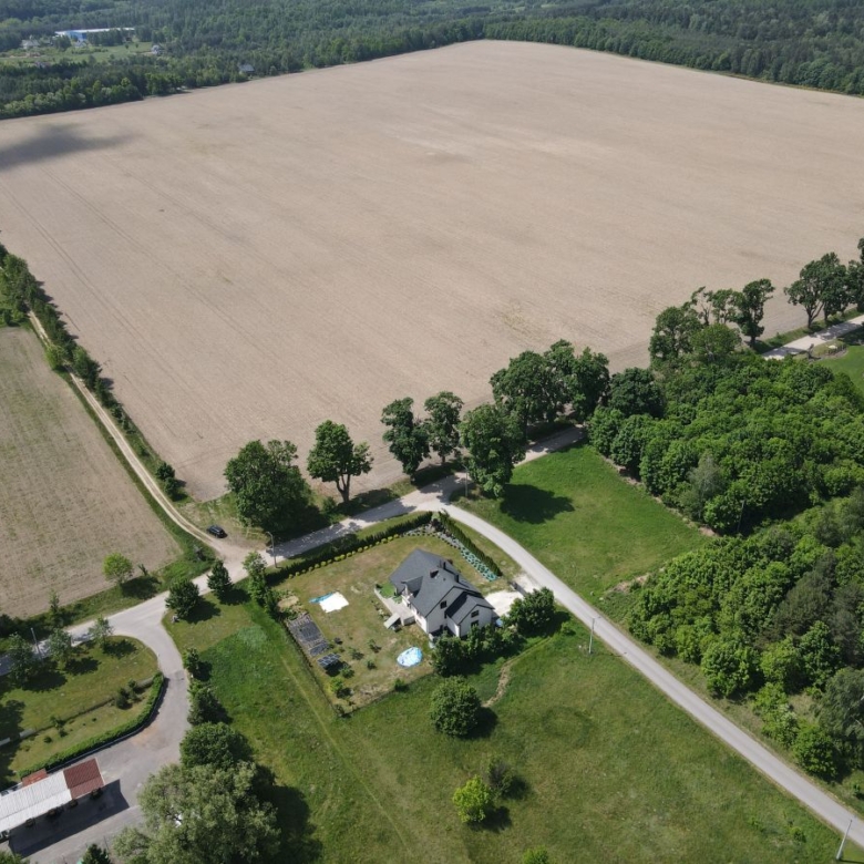 Aerial view of a rural property with a house, pool, and curved driveway along a tree-lined edge near fields and farmland.