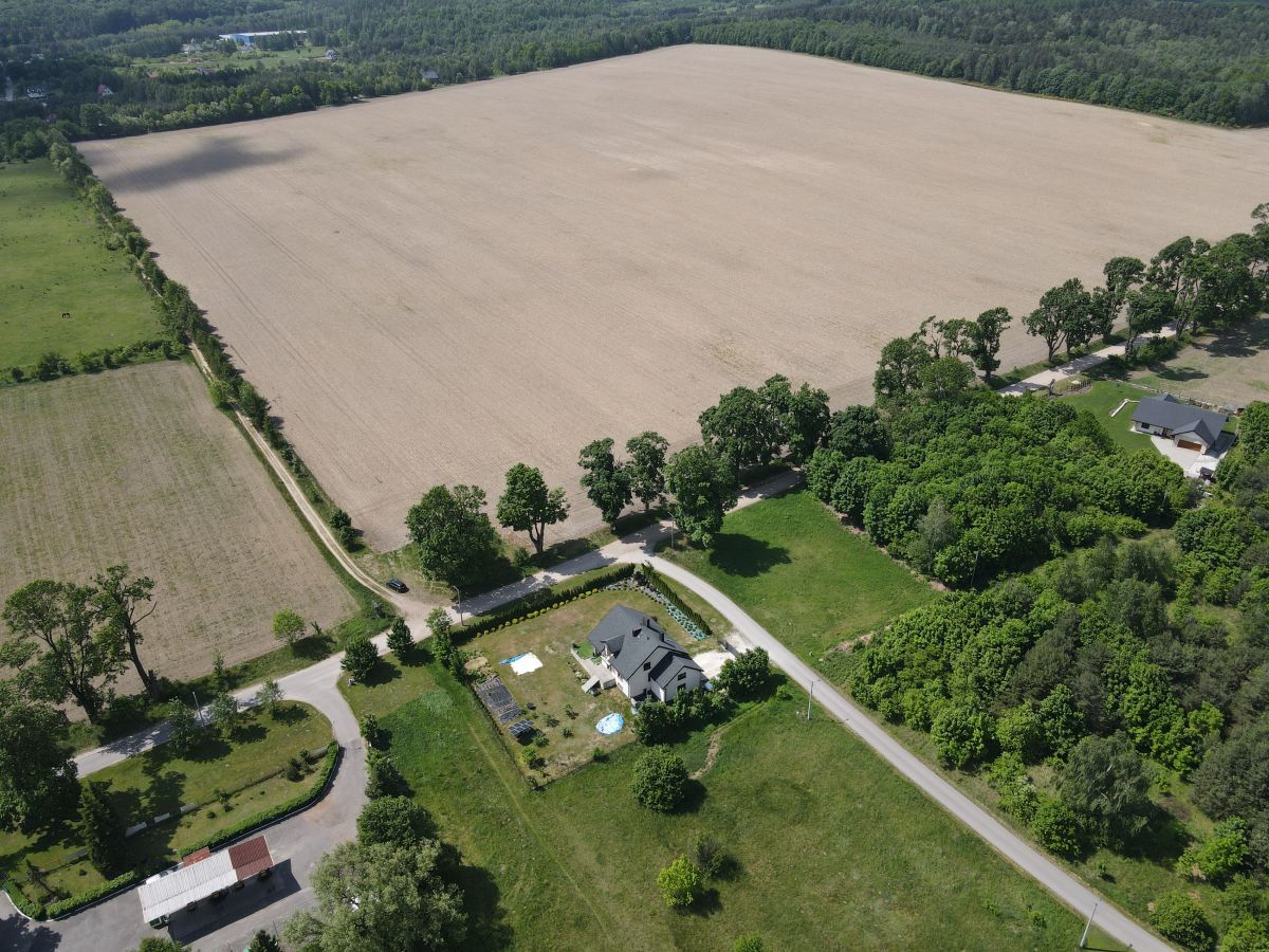 Aerial view of a rural property with a house, pool, and curved driveway along a tree-lined edge near fields and farmland.
