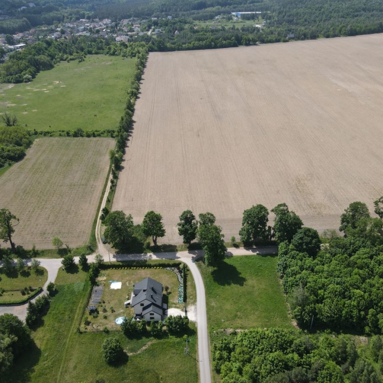 Aerial view of a rural landscape with a large plowed field on the right, a green meadow on the left, and a curving road passing a house and trees at the bottom center