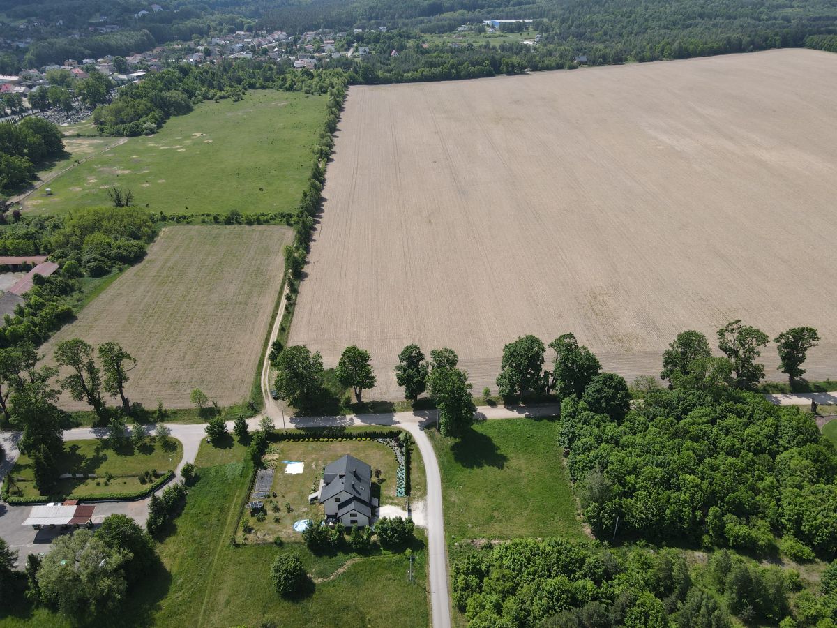 Aerial view of a rural landscape with a large plowed field on the right, a green meadow on the left, and a curving road passing a house and trees at the bottom center