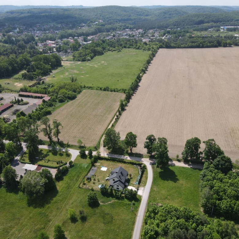 Aerial view of a rural landscape featuring a central house along a curved road, surrounding green fields and a large plowed field, with forests and a distant village at the horizon.