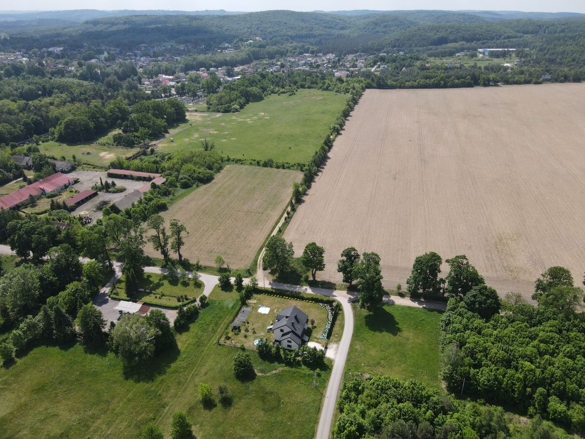 Aerial view of a rural landscape featuring a central house along a curved road, surrounding green fields and a large plowed field, with forests and a distant village at the horizon.