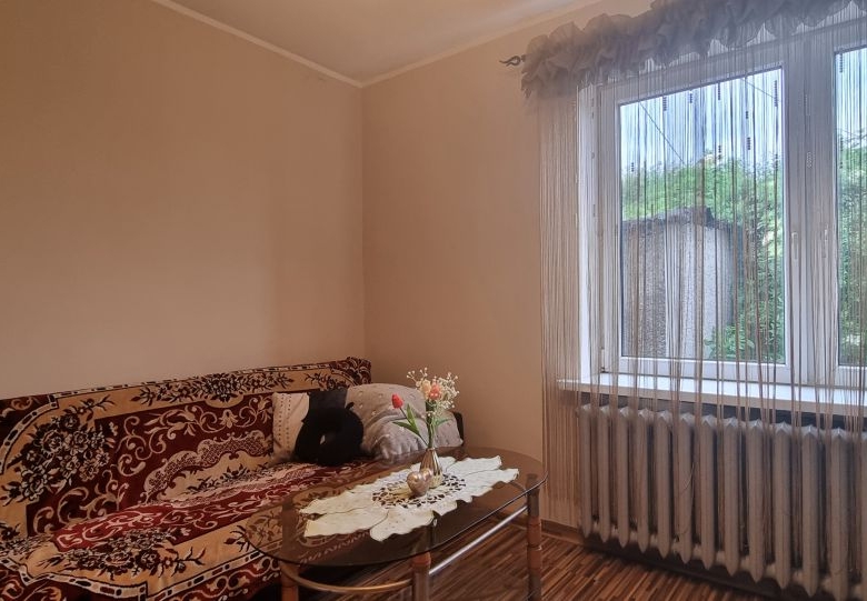 Cozy living room with a patterned sofa, glass coffee table, and a vase of flowers in front of a window with sheer curtains and a radiator beneath it.