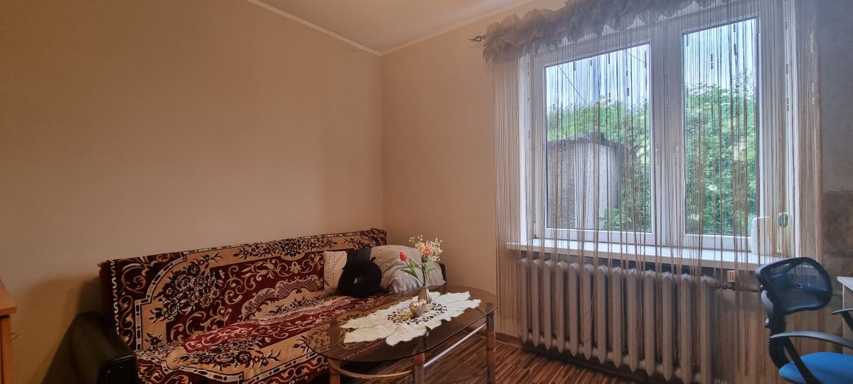 Cozy living room with a patterned sofa, glass coffee table, and a vase of flowers in front of a window with sheer curtains and a radiator beneath it.