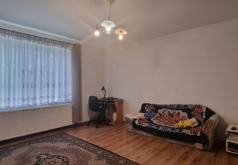 Living room with a large window dressed in sheer curtains, a small desk and chair in the corner, and a sofa covered with a floral blanket.