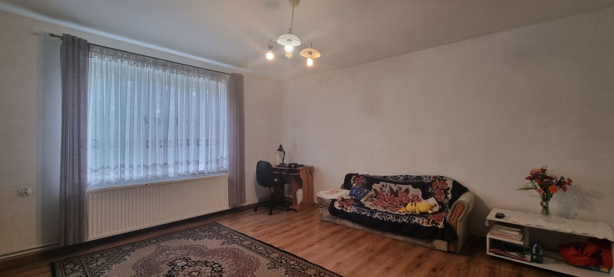 Living room with a large window dressed in sheer curtains, a small desk and chair in the corner, and a sofa covered with a floral blanket.