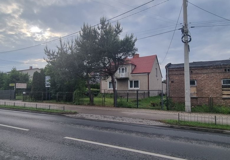 Residential street view with a beige house and a brick building beside a grassy yard, under cloudy skies and power lines.