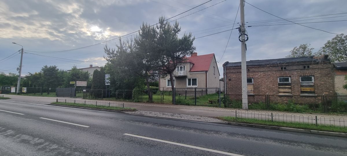 Residential street view with a beige house and a brick building beside a grassy yard, under cloudy skies and power lines.