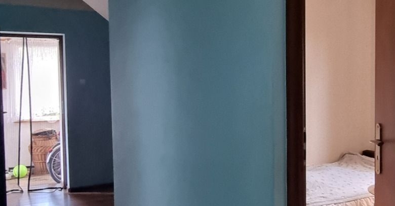 Blue-painted hallway wall with a doorway to a bedroom on the right; a left room shows a window, a bicycle wheel, and a green ball on the floor.