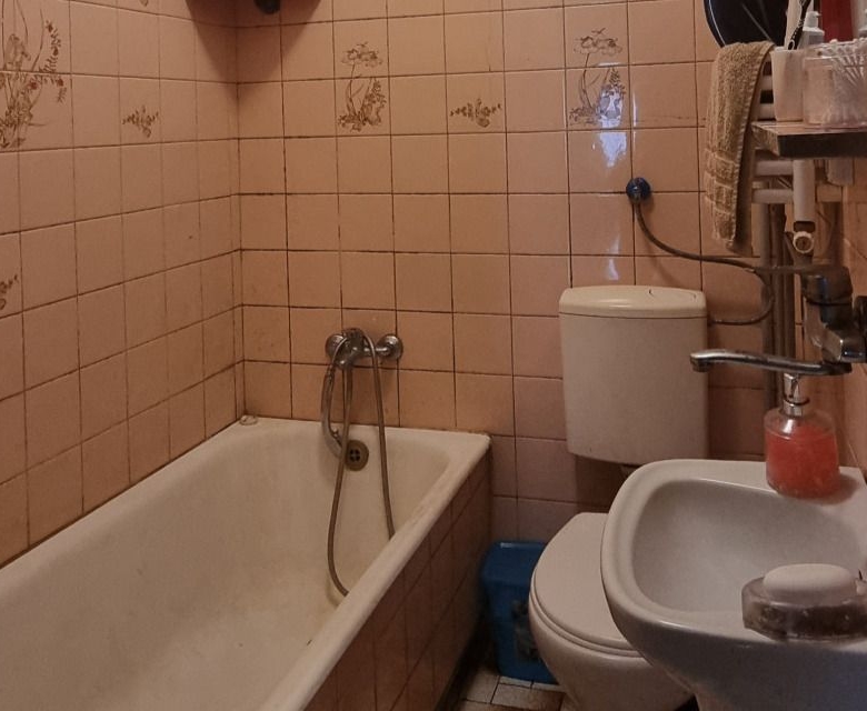 Narrow pink-tiled bathroom with a white bathtub, a toilet, and a small sink; a soap dispenser sits on the sink and towels are on a rack above the toilet area.