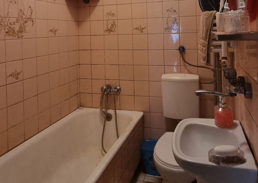 Narrow pink-tiled bathroom with a white bathtub, a toilet, and a small sink; a soap dispenser sits on the sink and towels are on a rack above the toilet area.