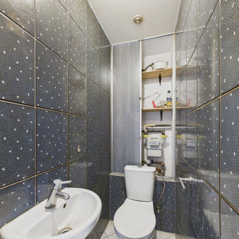 Narrow bathroom with blue mosaic tile walls, white toilet and sink, and a small shelf area with exposed plumbing behind the toilet.