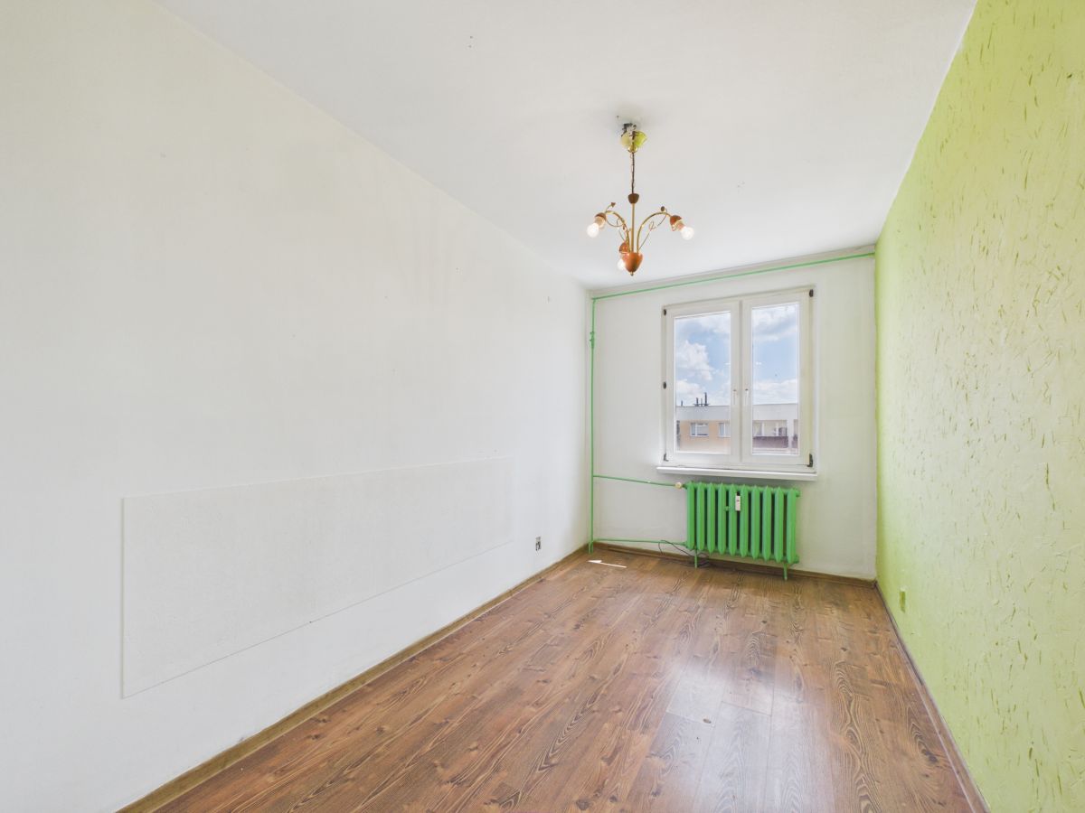 A nearly empty room with white walls, a lime-green accent wall, and a green radiator under the window; wooden floor and a hanging chandelier.