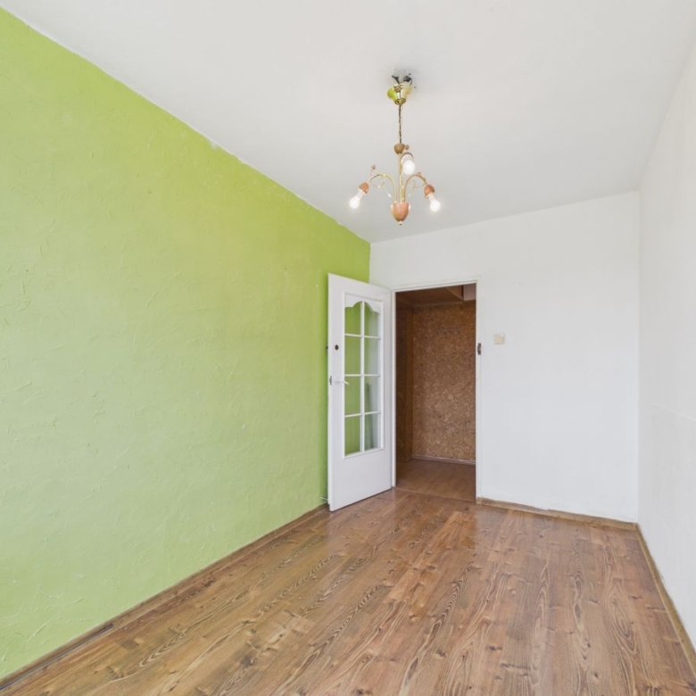 Empty room featuring a lime-green accent wall, white walls, and wooden flooring; an open door reveals another room and a vintage chandelier hangs from the ceiling.