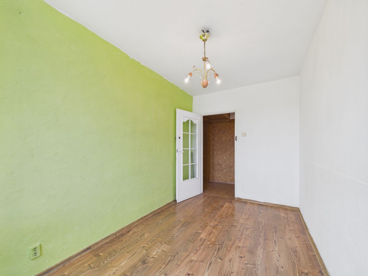 Empty room featuring a lime-green accent wall, white walls, and wooden flooring; an open door reveals another room and a vintage chandelier hangs from the ceiling.