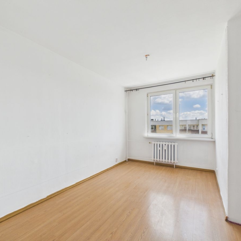 Empty white-walled room with light wood laminate flooring and a radiator under a large window overlooking neighboring buildings.