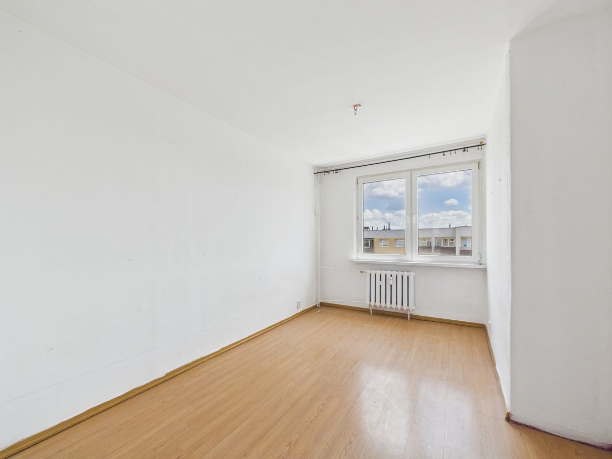 Empty white-walled room with light wood laminate flooring and a radiator under a large window overlooking neighboring buildings.
