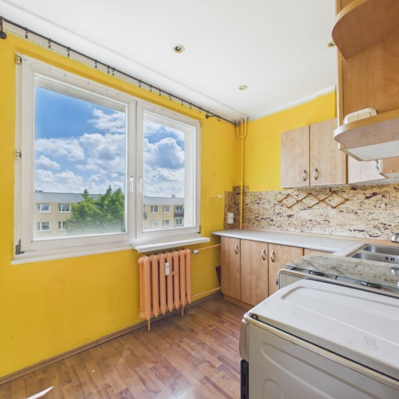 Bright yellow kitchen with a large window showing a blue sky and apartment buildings, radiator below the window, and wooden cabinets along the wall.