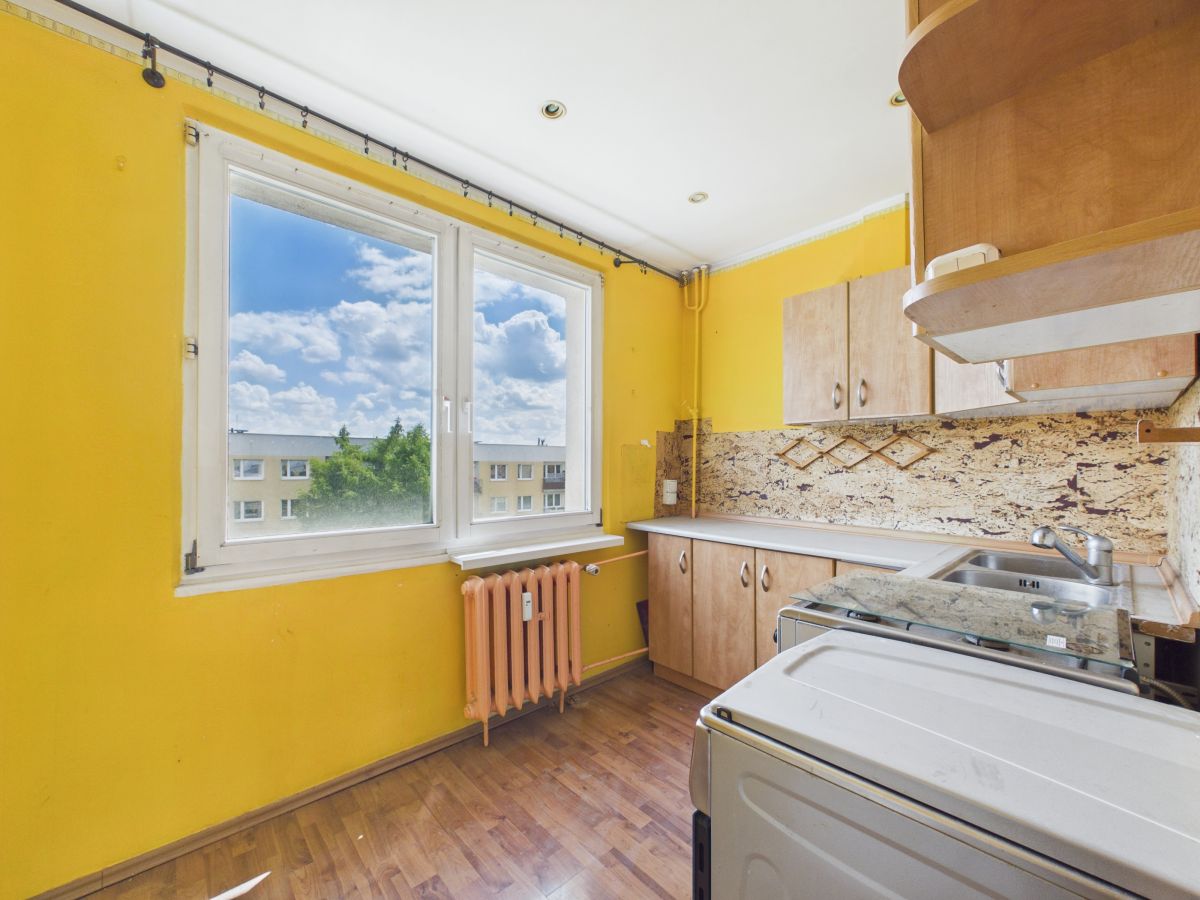 Bright yellow kitchen with a large window showing a blue sky and apartment buildings, radiator below the window, and wooden cabinets along the wall.