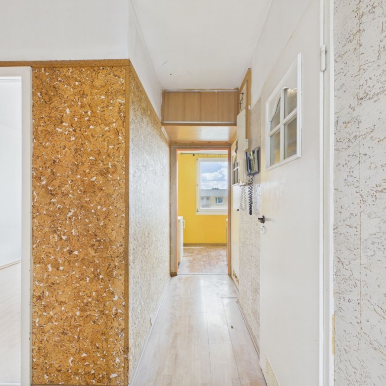 Narrow hallway in a retro apartment with cork walls, white doors, and a bright yellow room visible at the far end through an open door.