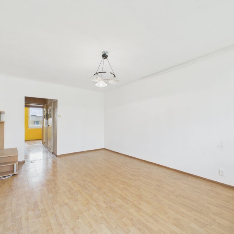 Empty living room with wooden floor, white walls, and a doorway to a yellow kitchen; a small wooden shelf on the left.