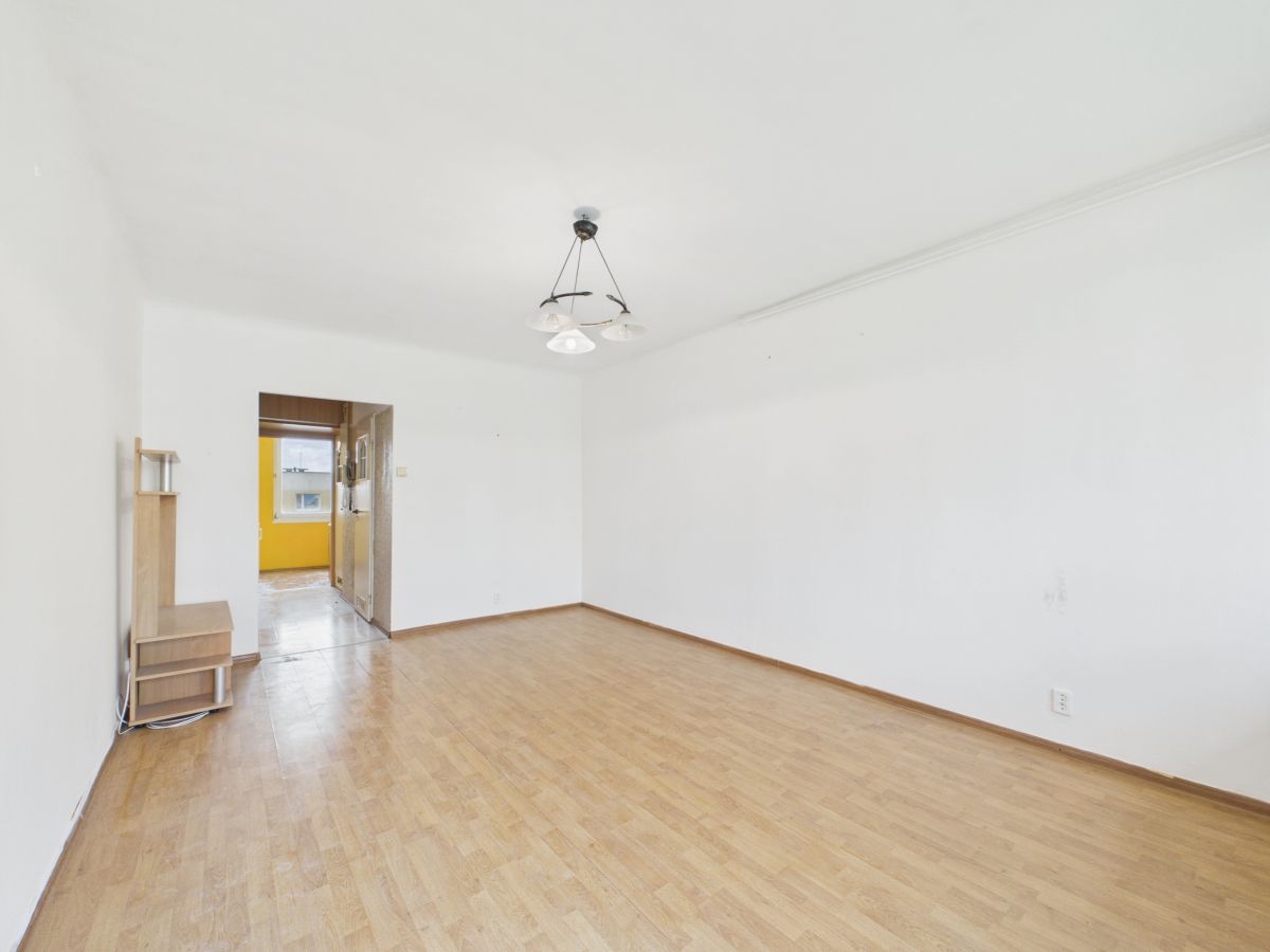 Empty living room with wooden floor, white walls, and a doorway to a yellow kitchen; a small wooden shelf on the left.