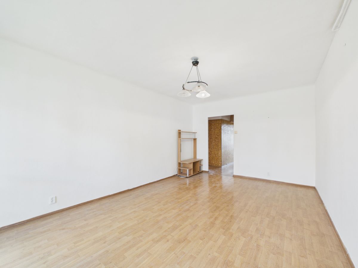 Empty living room with light wood floor and white walls, a small wooden shelf in the corner, and a doorway to a brown textured hallway; ceiling light fixture above