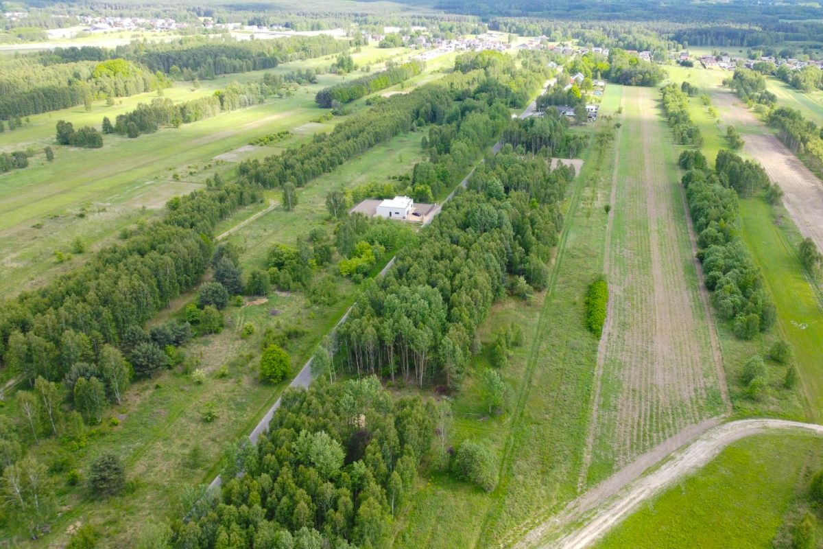 Aerial view of a rural landscape with a central tree-lined road, fields, and a white building surrounded by trees.