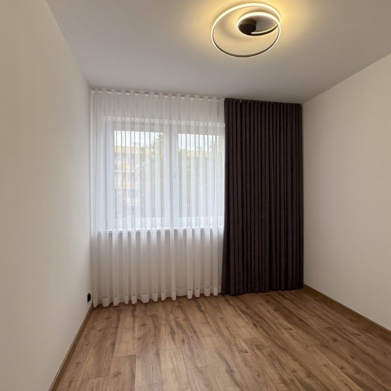 Empty room with light wood floor, white walls, and a large window with sheer white curtains and a dark drape.