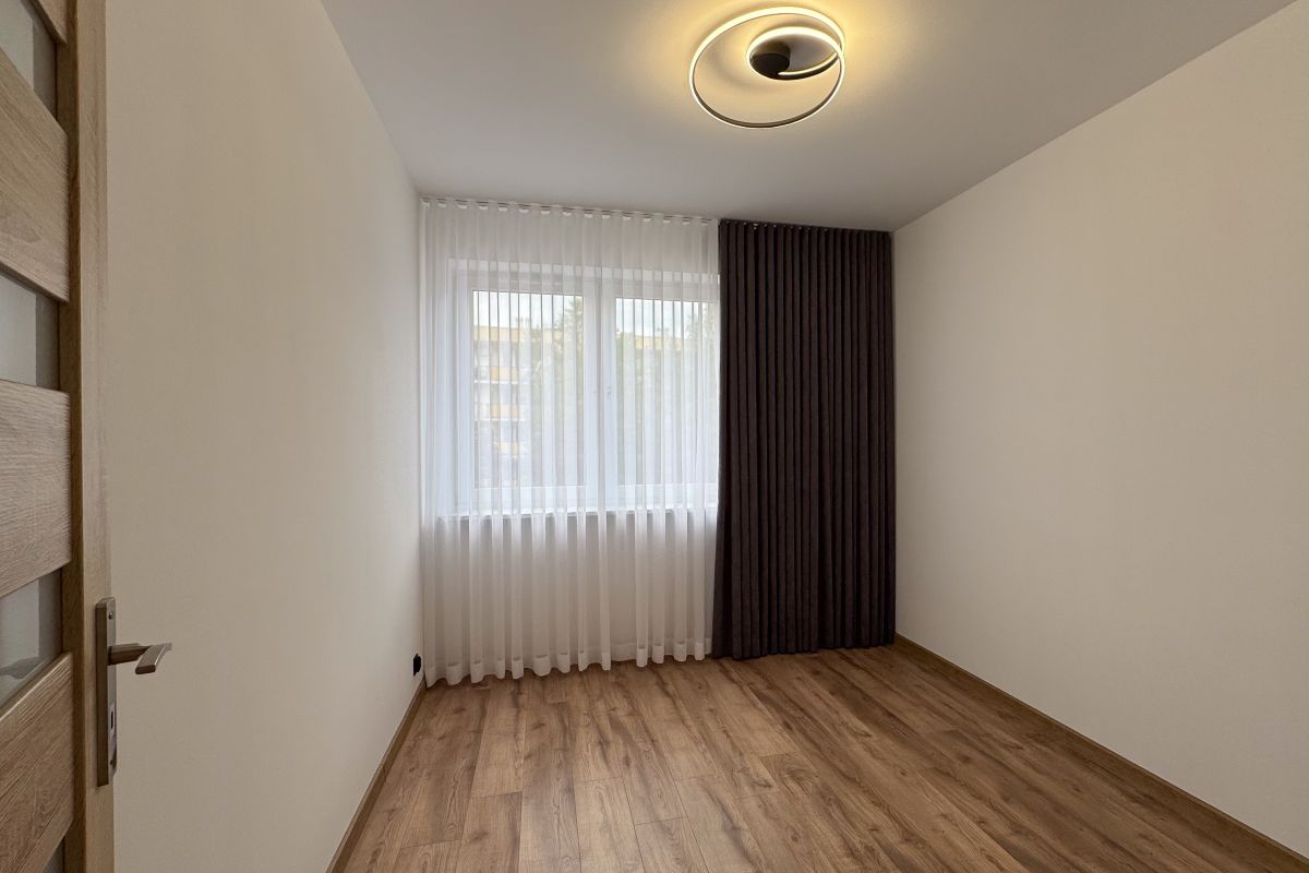 Empty room with light wood floor, white walls, and a large window with sheer white curtains and a dark drape.