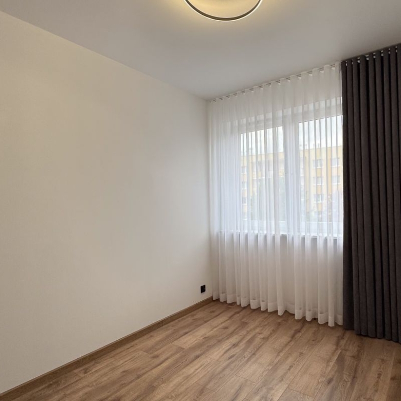 Empty modern room with light wood flooring, white walls, and a large window with sheer white curtains and dark drapes on the right edge.