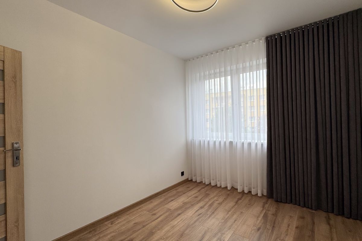 Empty modern room with light wood flooring, white walls, and a large window with sheer white curtains and dark drapes on the right edge.