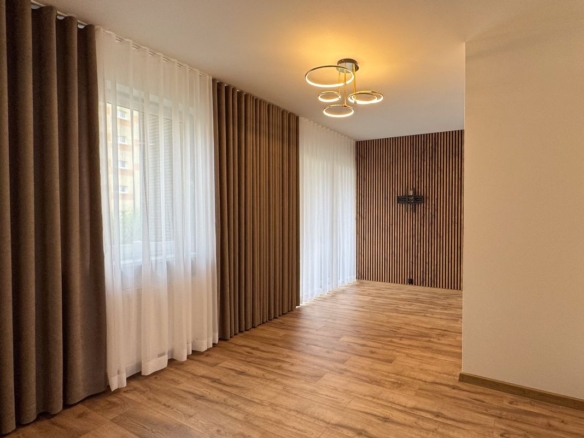 Empty living room with wood slat accent wall, large windows with sheer white and brown curtains, and a circular ceiling light.