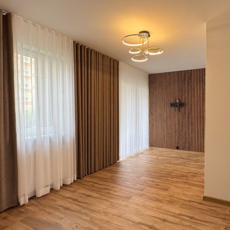 Empty living room with wood slat accent wall, large windows with sheer white and brown curtains, and a circular ceiling light.