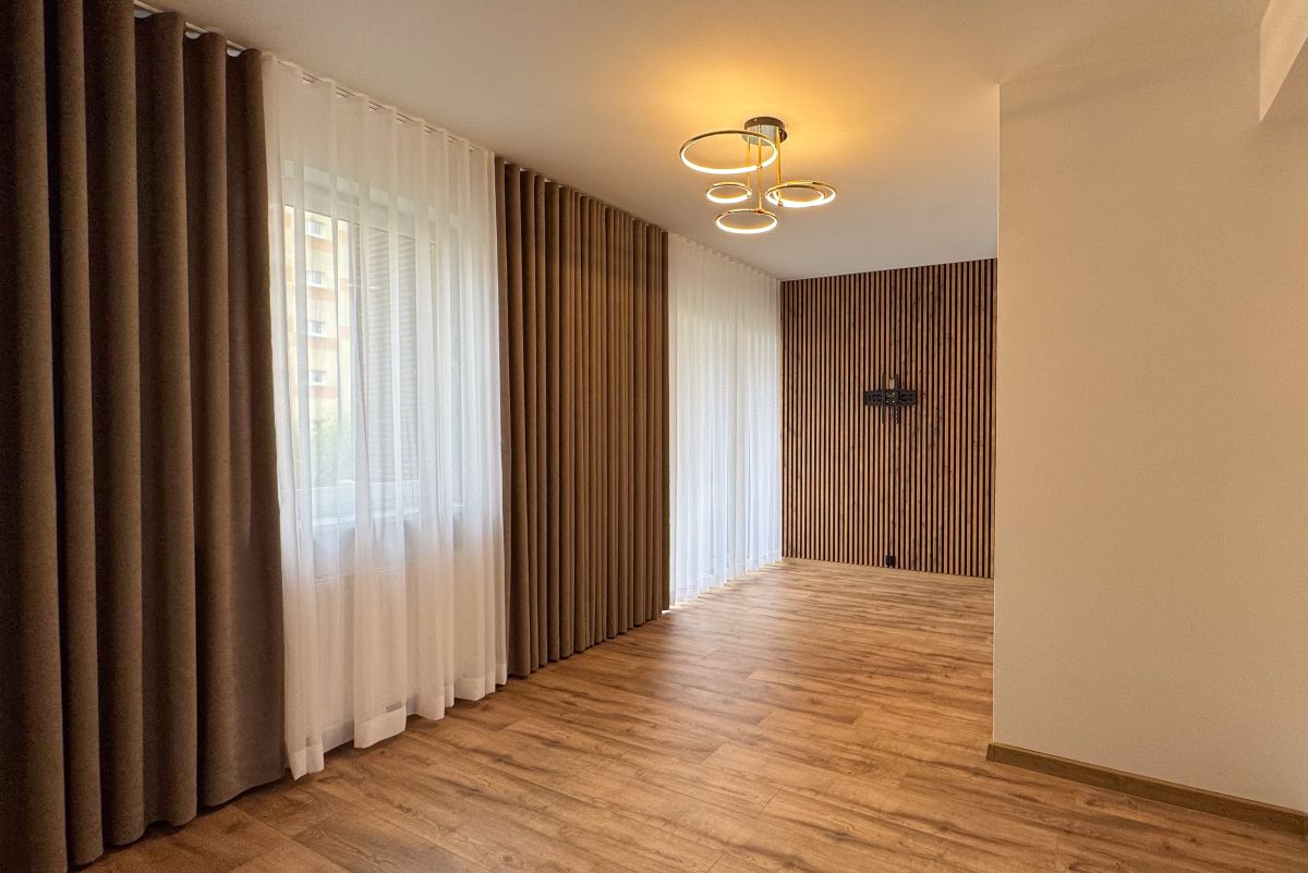 108854807.jpg Empty living room with wood slat accent wall, large windows with sheer white and brown curtains, and a circular ceiling light.